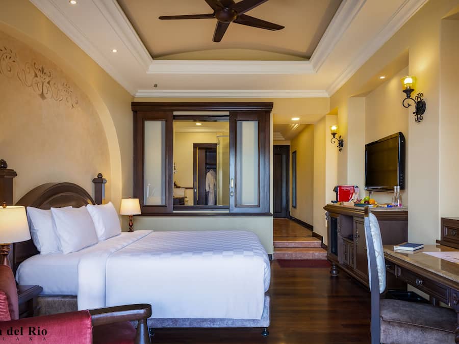 A hotel room with a large bed, a desk with a chair, a TV, and a ceiling fan.