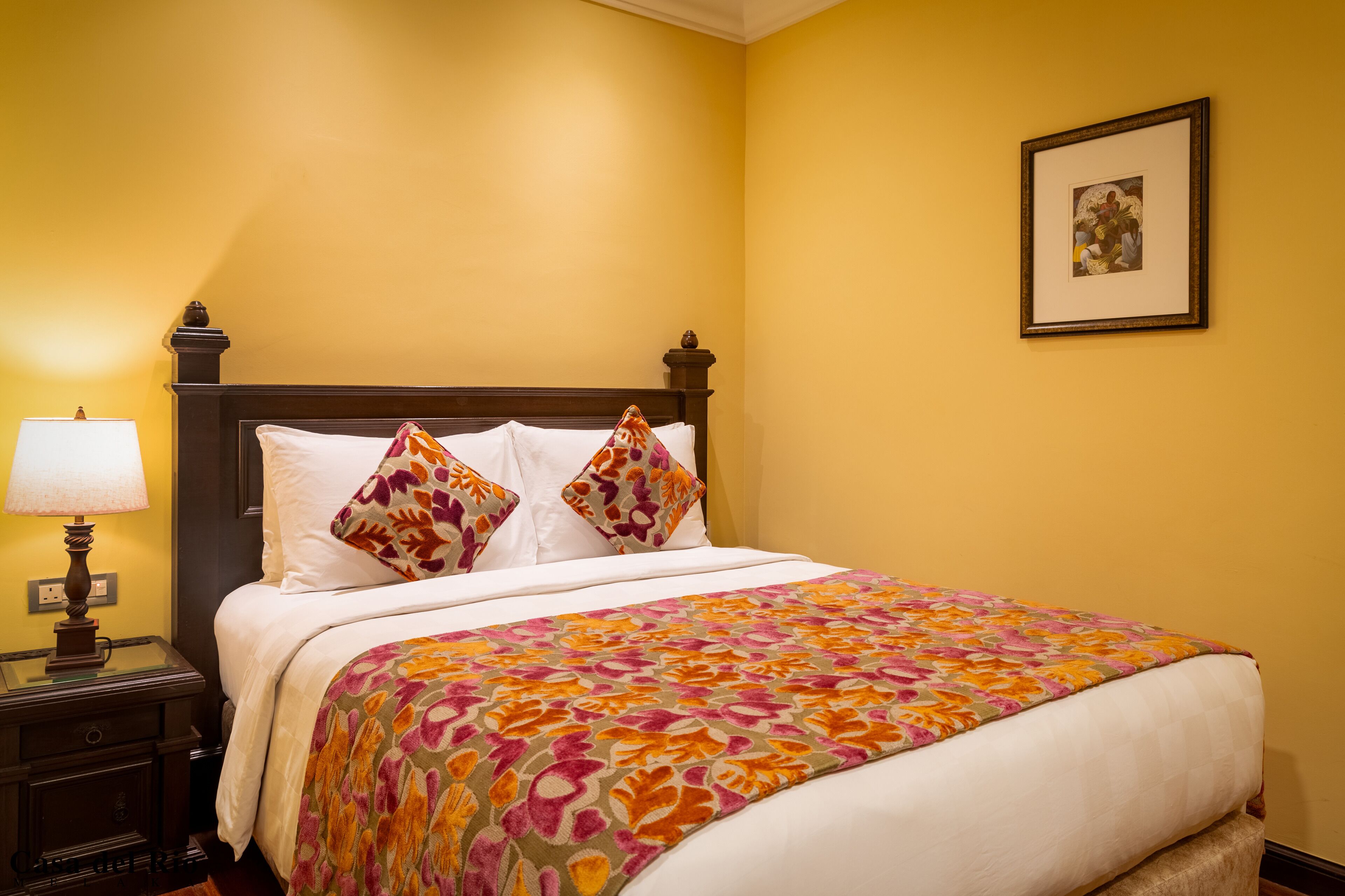 A neatly made bed with a floral bedspread, a bedside lamp, and a framed picture on the wall.