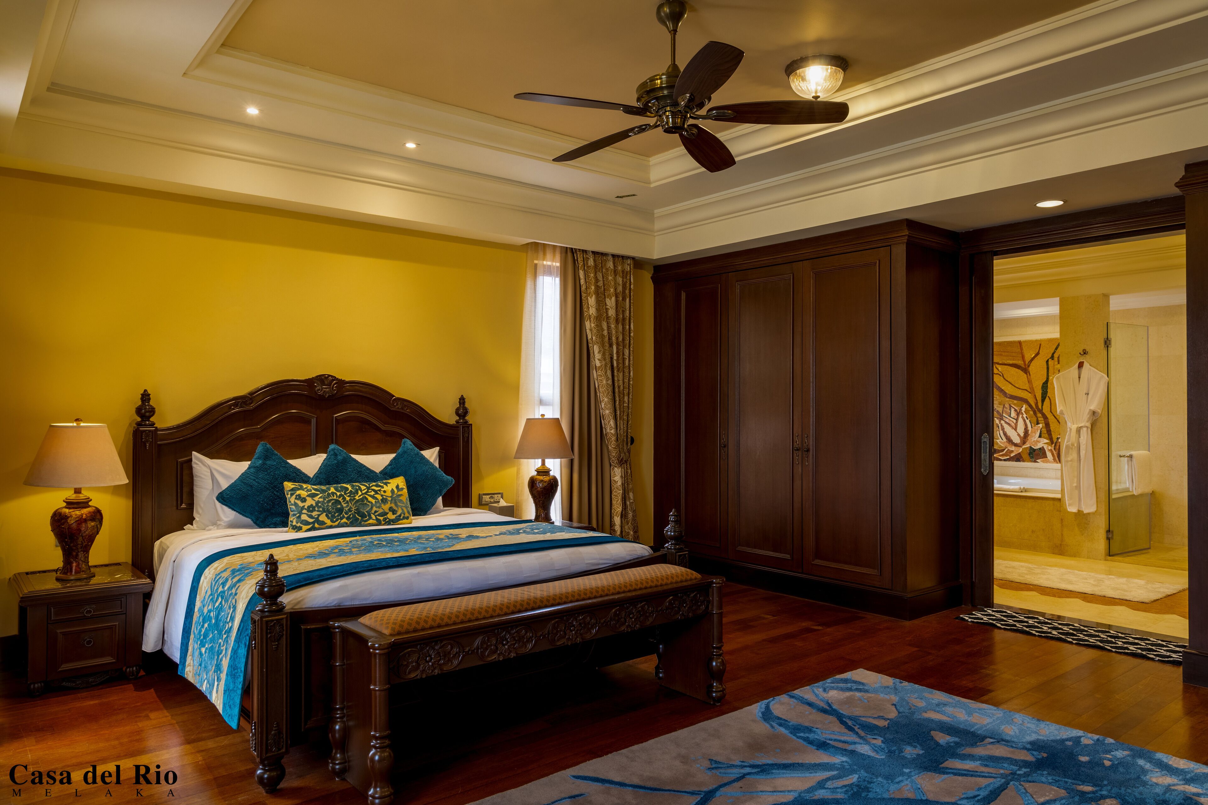 A hotel room with a large bed, wooden headboard, two bedside lamps, a ceiling fan, and a view of a bathroom with a glass door.