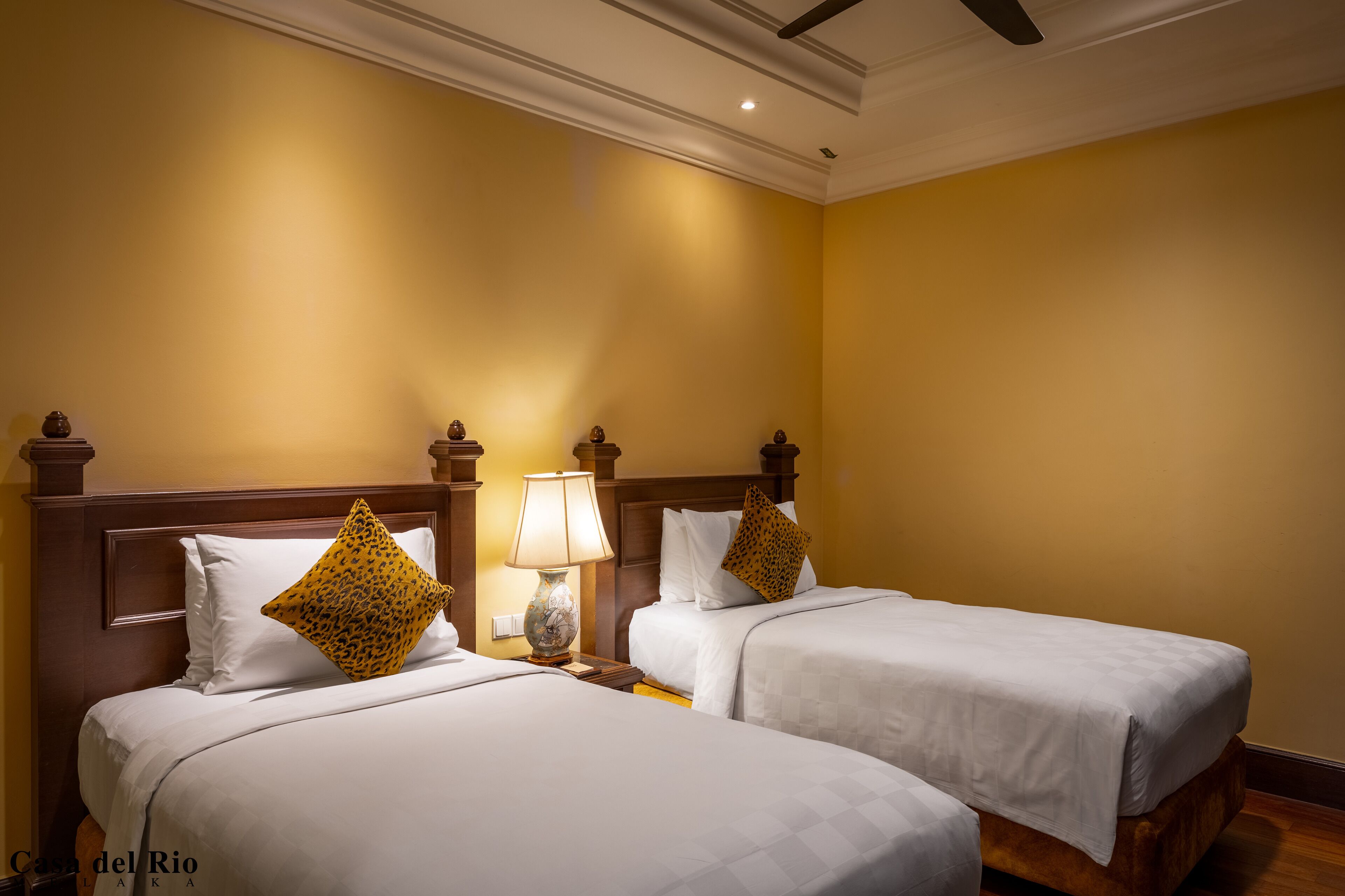 A hotel room with two beds, each with white bedding and a wooden headboard. There is a nightstand with a lamp between the beds.