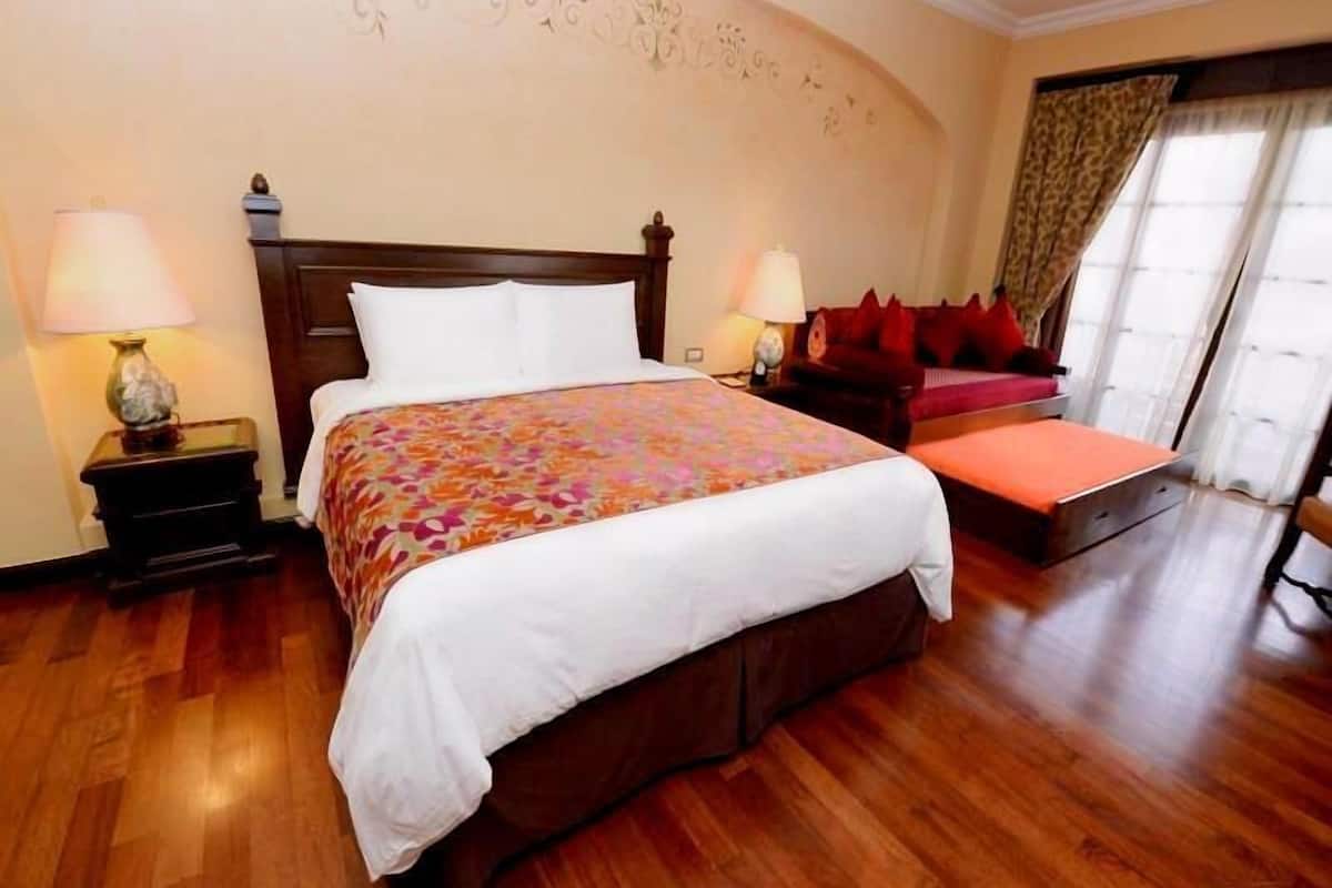 A hotel room with a large bed, two bedside tables with lamps, a red sofa, and a window with curtains.