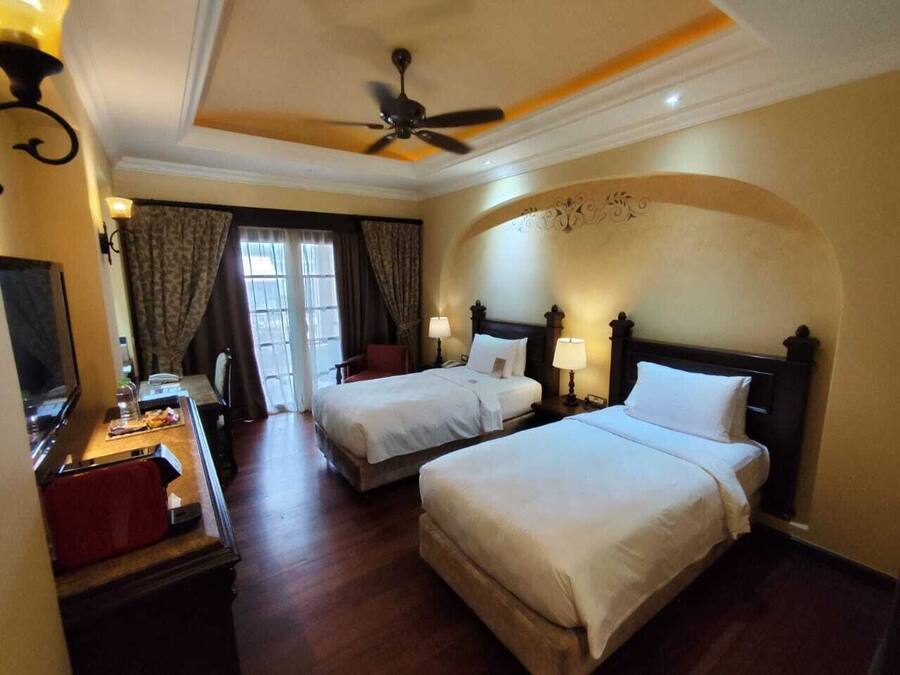 A hotel room with two beds, a desk with a chair, a television, and a ceiling fan.