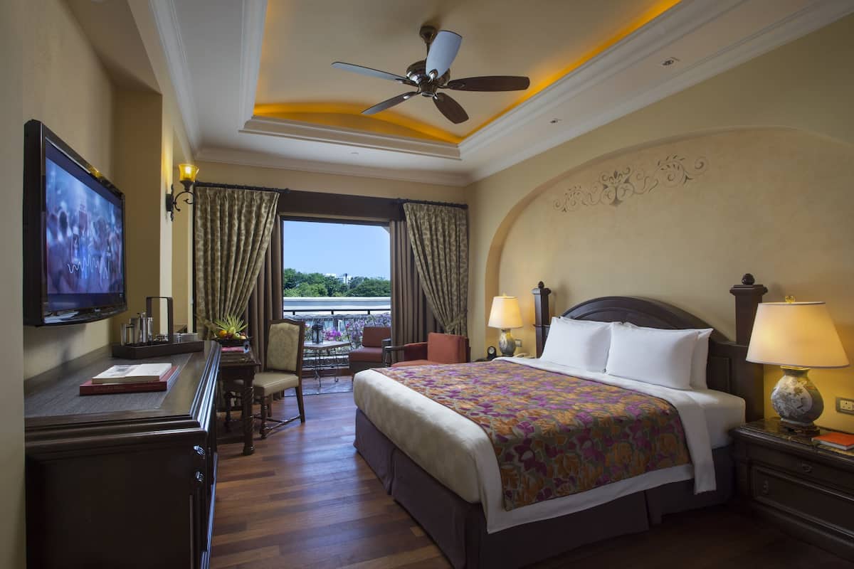 A hotel room with a bed, a desk with a TV, a chair, a lamp, and a view of the outdoors.
