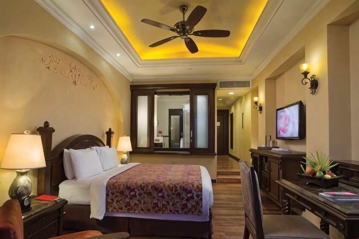 Habitación de hotel con cama, lámparas de noche, escritorio, silla, televisor y ventilador de techo.