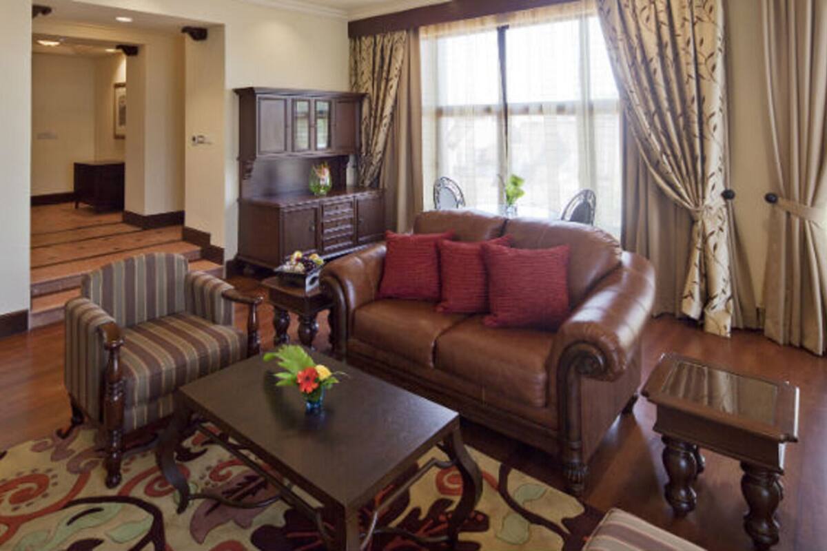 A living room with a brown leather sofa, striped armchair, and a wooden coffee table with a vase. A dark wooden cabinet is against the wall, and a window with curtains is visible.