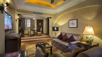 Uma sala de estar com sofá, poltronas, mesa de centro e televisão. Ao fundo, há uma área de jantar com mesa e cadeiras.
