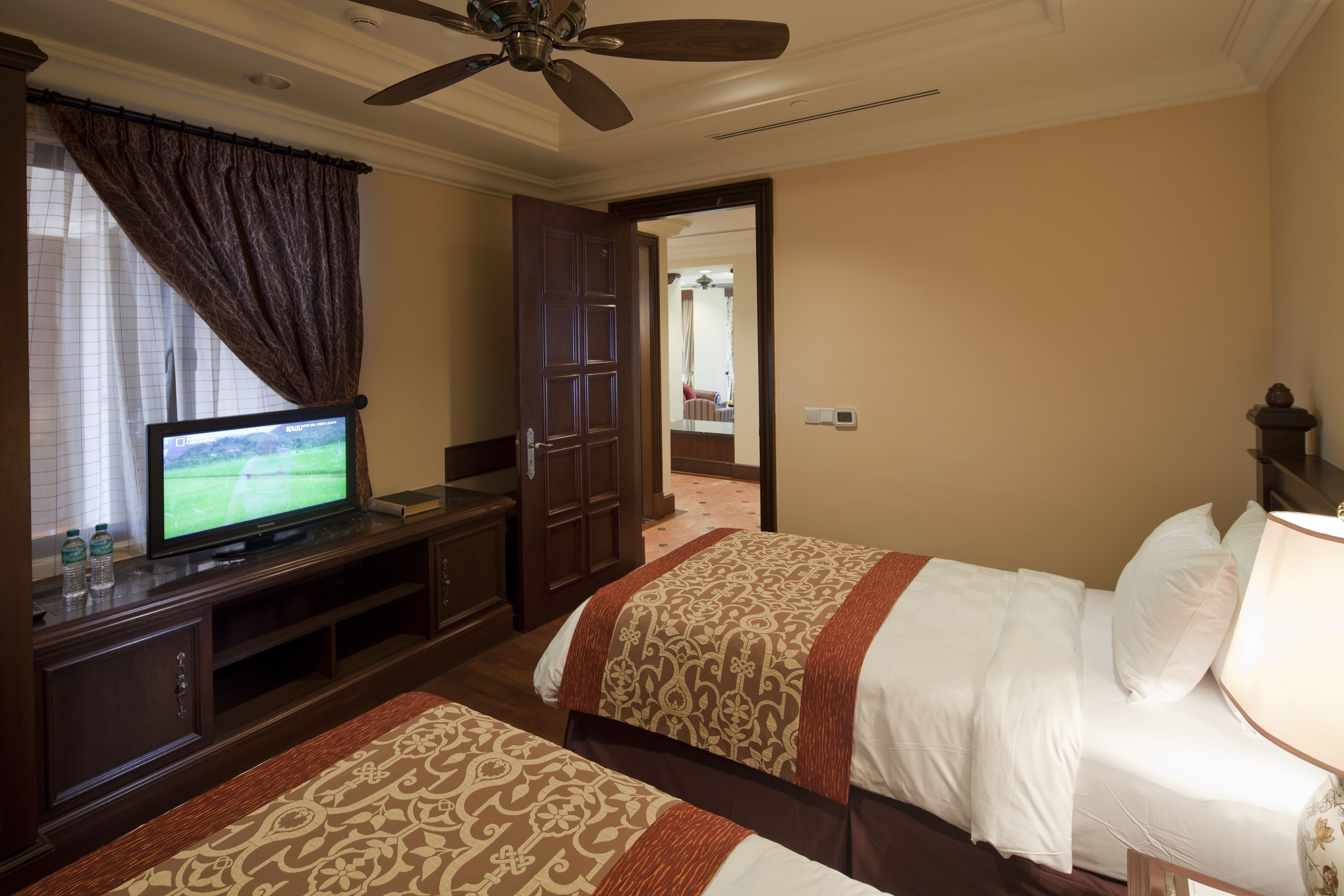 Habitación de hotel con televisor, armario, dos camas con cubrecamas estampados, ventilador de techo y vista hacia la habitación contigua.