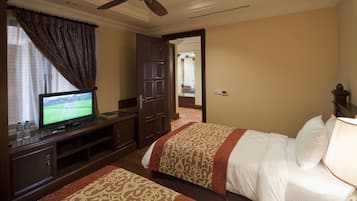 Habitación de hotel con televisor, armario, dos camas con cubrecamas estampados, ventilador de techo y vista hacia la habitación contigua.