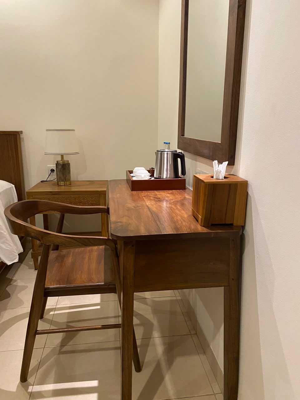 A hotel room with a wooden desk, a chair, a bedside table with a lamp, a coffee maker, and a tissue box.