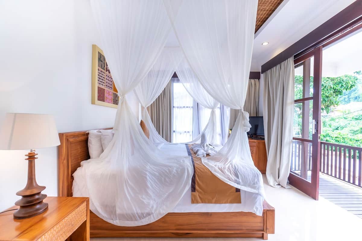 Un dormitorio con cama con dosel, una mesita de noche con lámpara y vistas al exterior con vegetación.