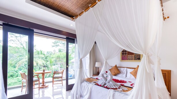 A spacious bedroom with a canopy bed, a balcony with wooden chairs, and a view of lush greenery. - Villa Sonia Bisma (Ubud)