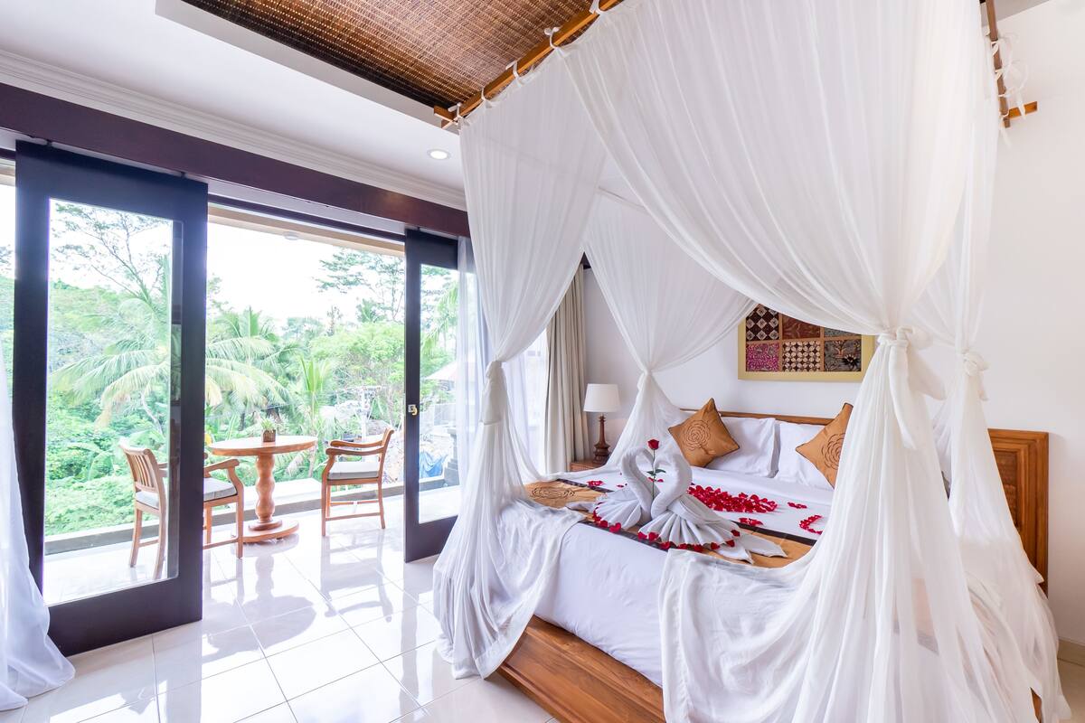 Un dormitorio amplio con cama con dosel, un balcón con sillas de madera y vistas a exuberante vegetación.