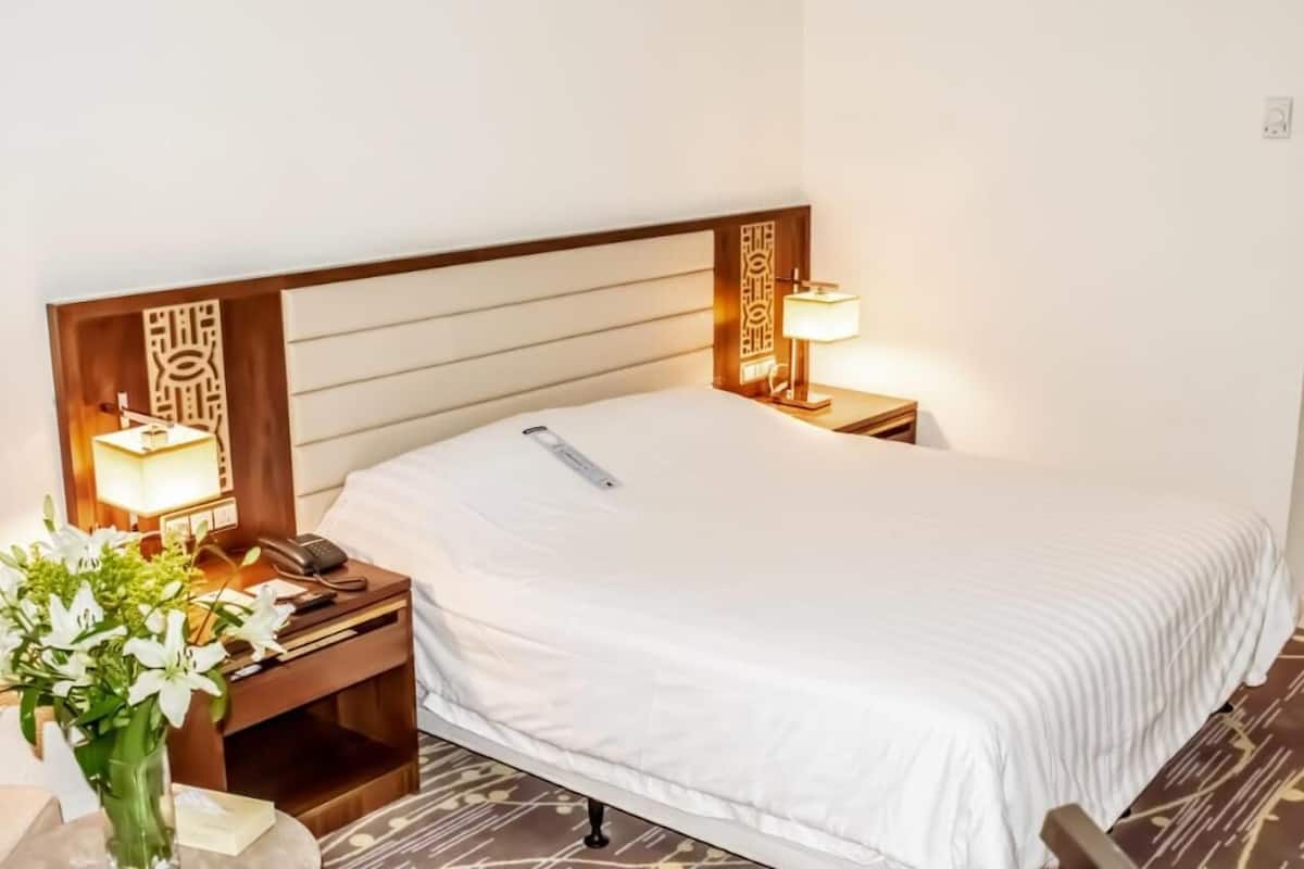 A hotel room with a neatly made bed, a bedside table with a lamp, a phone, and a vase with flowers.