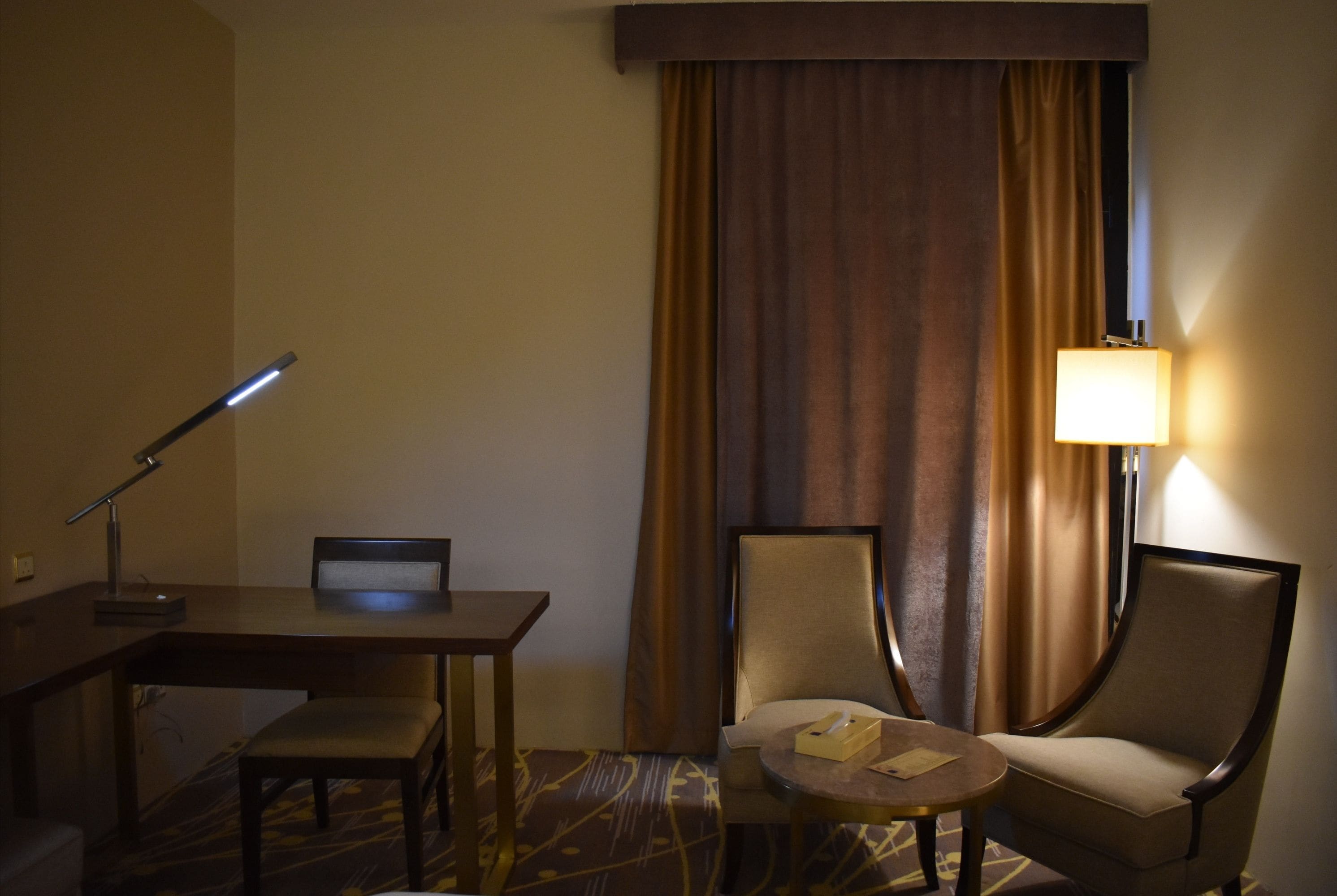 A hotel room with a desk, two chairs, a lamp, and a bed.