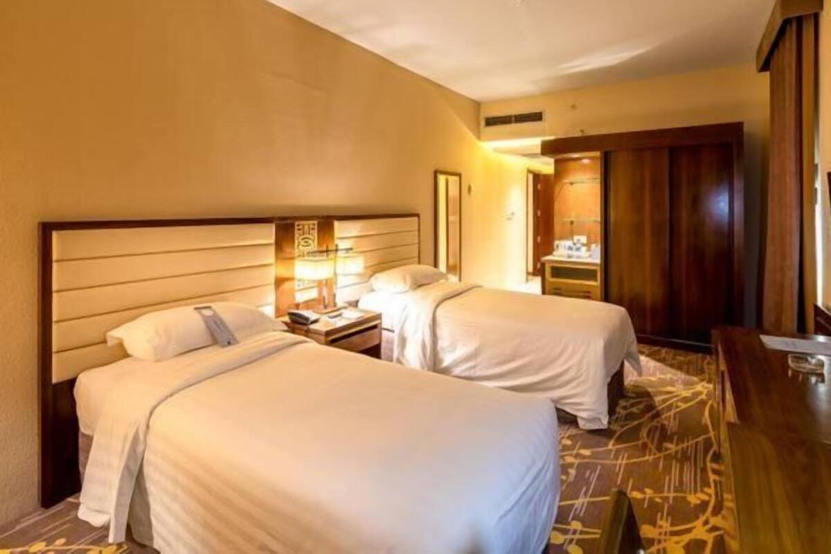 A hotel room with two beds, a wooden headboard, a nightstand with a lamp, and a wooden desk.