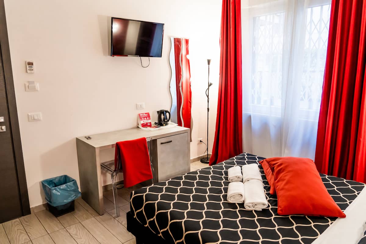 A hotel room with a bed, a desk, a TV, and a window with red curtains.