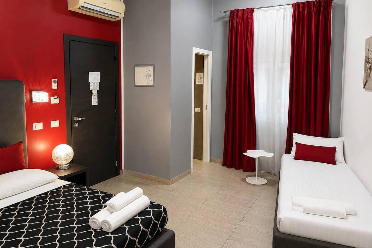 A hotel room with a bed, a bedside table with a lamp, a small bedside table with a lamp, a window with red curtains, and an air conditioning unit on the wall.
