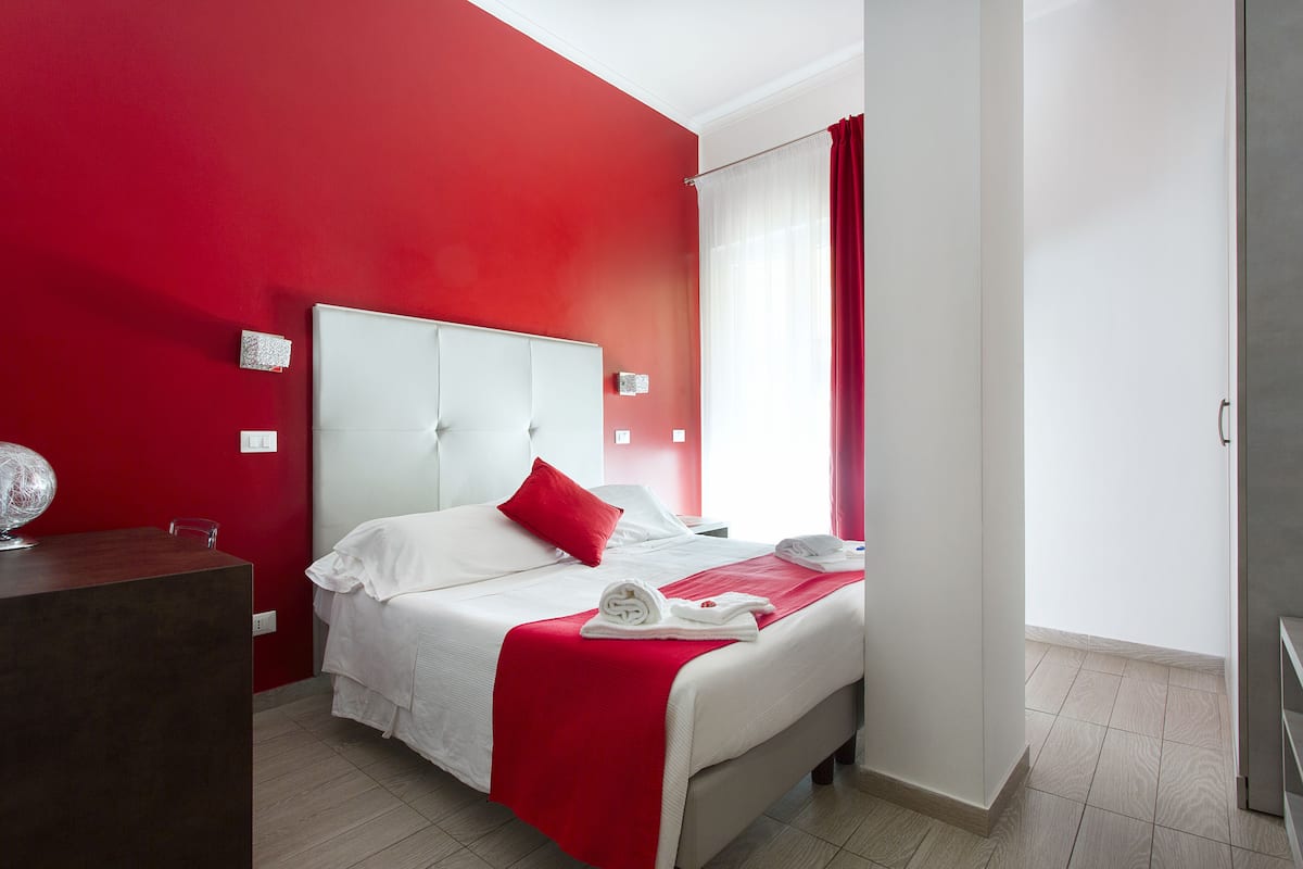 A hotel room with a red accent wall, a white bed with red pillows, a desk with a globe, and a bathroom visible through a door.