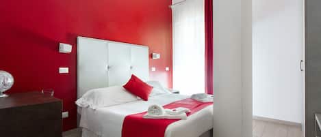 A hotel room with a red accent wall, a white bed with red pillows, a desk with a globe, and a bathroom visible through a door.