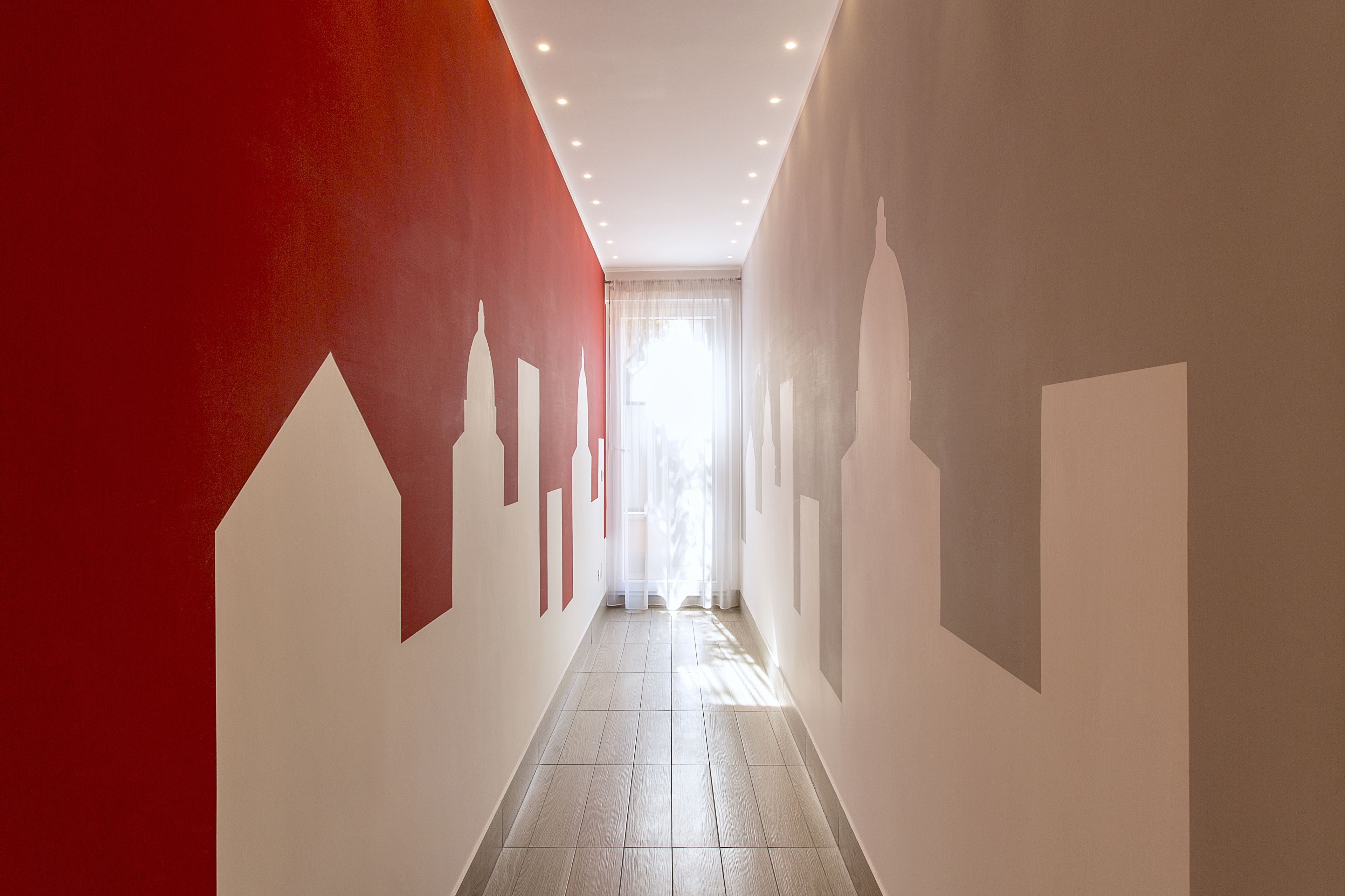 A hallway with a red and white color scheme, light wooden flooring, and a view of a bright room at the end.