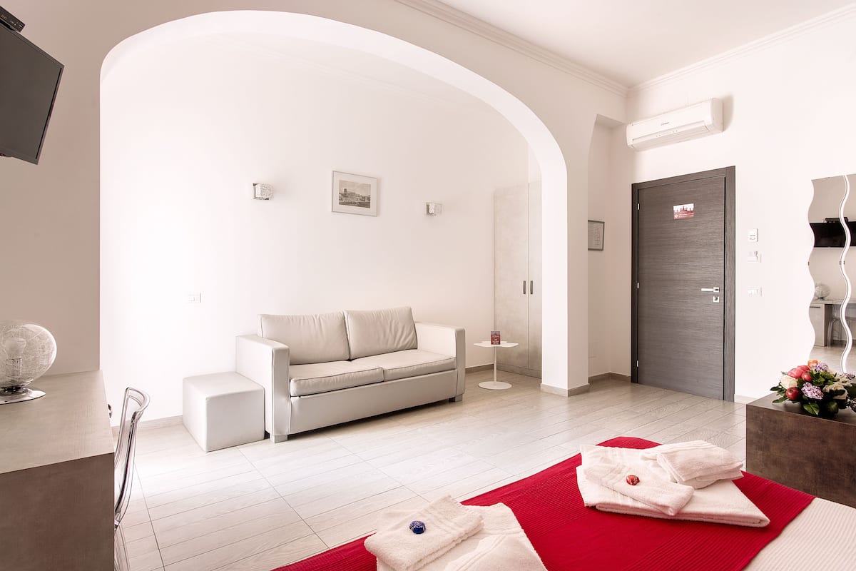A modern living room with a sofa, a small table, and a red rug. There is a door and a wall-mounted air conditioner.