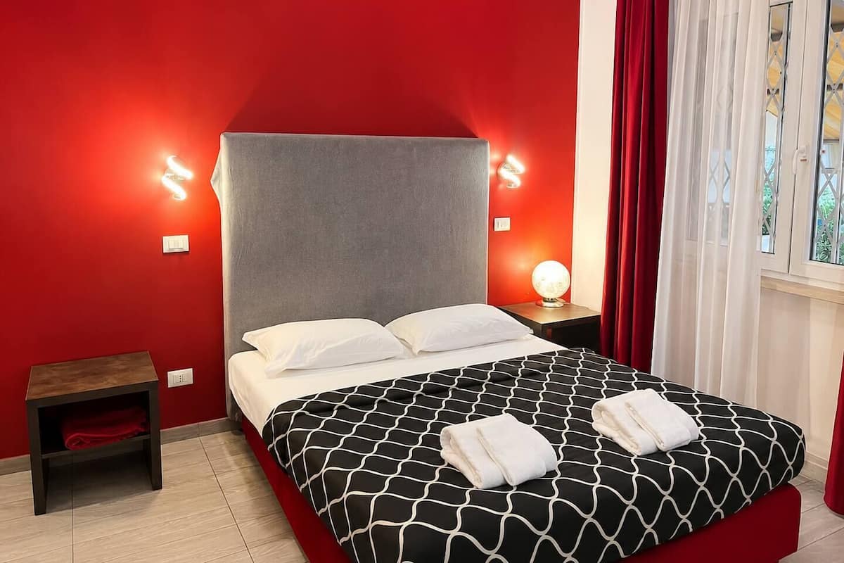 A bed with a grey headboard, white pillows, and a black and white patterned bedspread. There is a small wooden nightstand with a red towel and a round table lamp. The room has red walls and a window with white curtains.