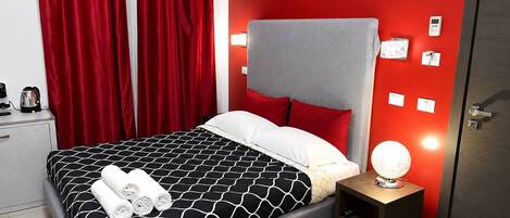 A modern hotel room with a bed, red curtains, a bedside table with a lamp, and a door.