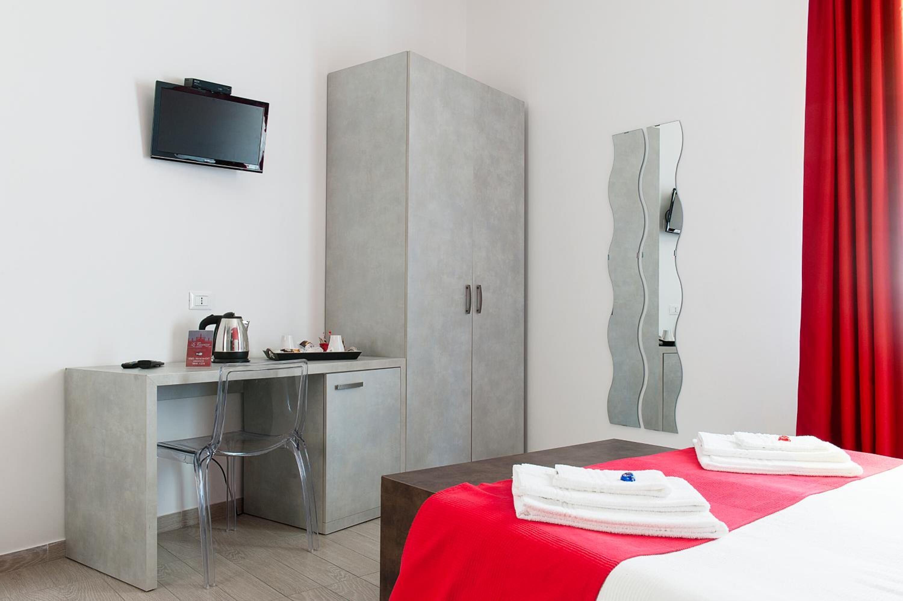 Et moderne hotelværelse med en grå garderobe, et lille skrivebord med elkedel, en stol, en seng med rødt sengetøj og et fjernsyn ophængt på væggen.