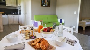 A breakfast table with croissants, fruit, and beverages. There is a green couch and a bed in the background.