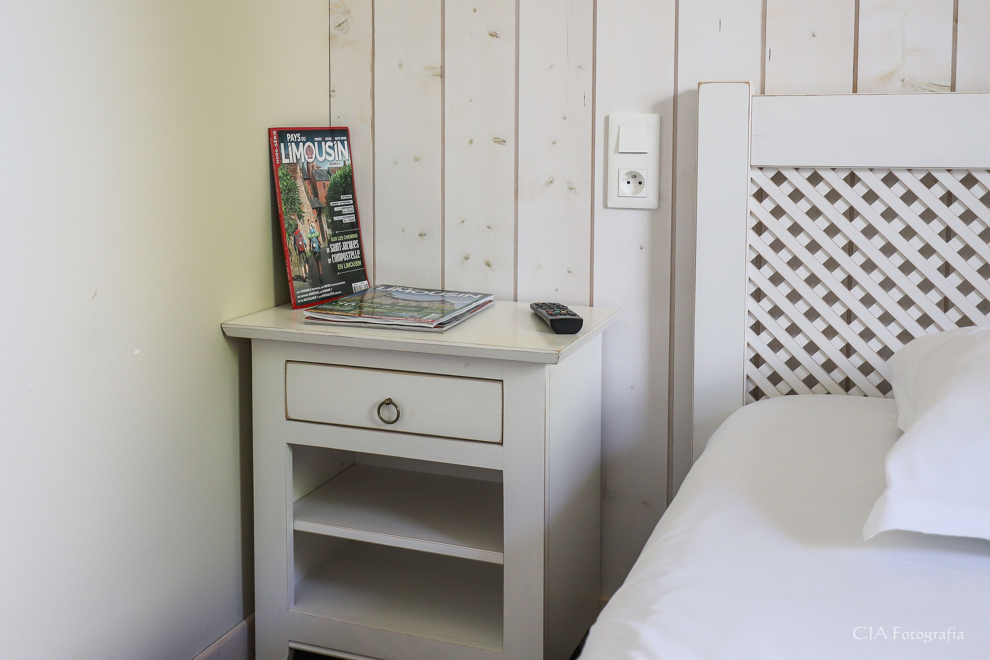 A hotel room with a white nightstand, a magazine, a remote control, and a bed with white bedding.