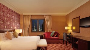 A hotel room with a large bed, a desk, a chair, a TV, and a view of a cityscape with two towers. - Dallah Taibah Hotel (Madinah)