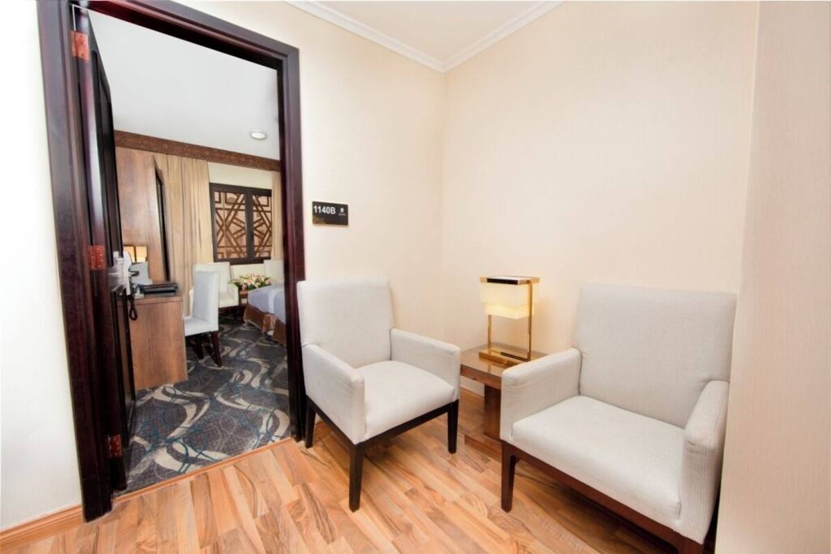 A hotel room with a wooden floor, two armchairs, a small table with a lamp, and a door leading to another room.