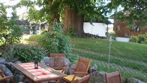 Un patio al aire libre con mesas y sillas de madera, rodeado de follaje y un muro de piedra.