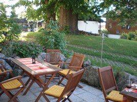 Un patio al aire libre con mesas y sillas de madera, rodeado de follaje y un muro de piedra.