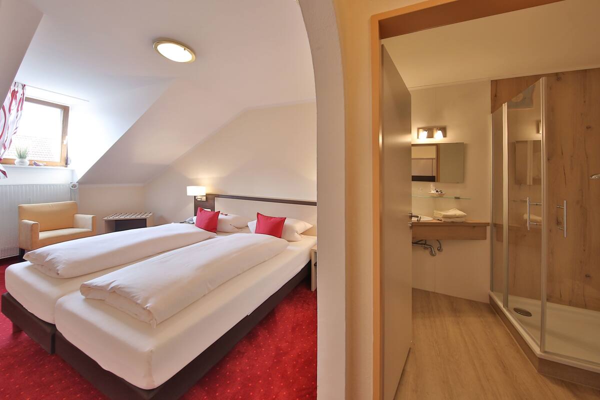 A hotel room with two beds, a red carpet, a wooden floor, a shower with glass doors, and a window with curtains.