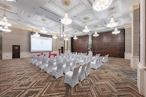 A conference room with rows of white chairs, a large screen, and ornate chandeliers.