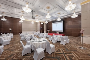 Un salón de conferencias con mesas redondas, sillas blancas, candelabros y una pantalla de proyección.