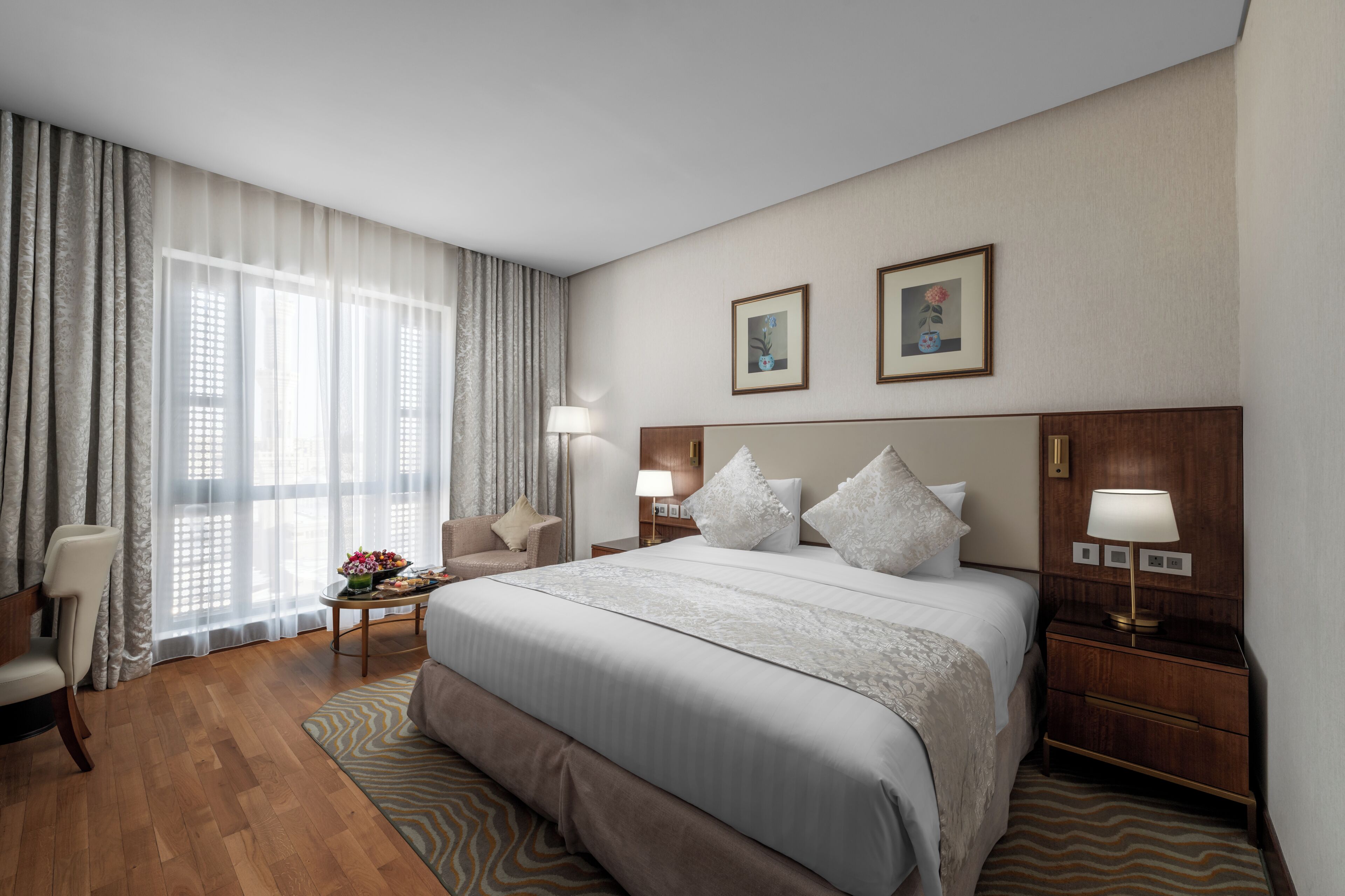 Habitación de hotel con una cama grande, un rincón de descanso con una silla, una mesita con un florero y dos cuadros en la pared.