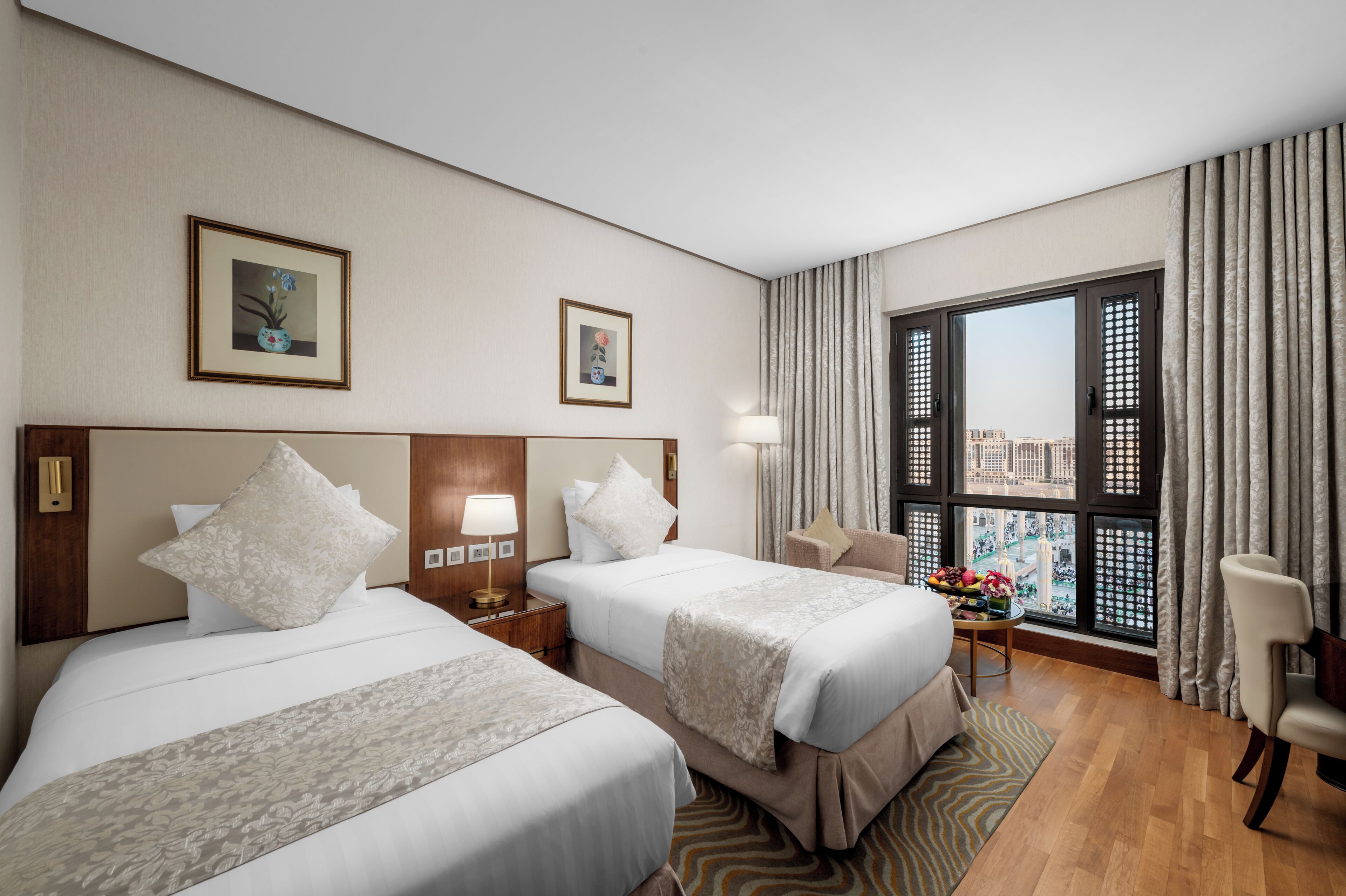 Habitación de hotel con dos camas, una mesita de noche, una lámpara, una silla y vistas de la ciudad a través de amplias ventanas.