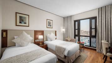 Habitación de hotel con dos camas, una mesita de noche, una lámpara, una silla y vistas de la ciudad a través de amplias ventanas.