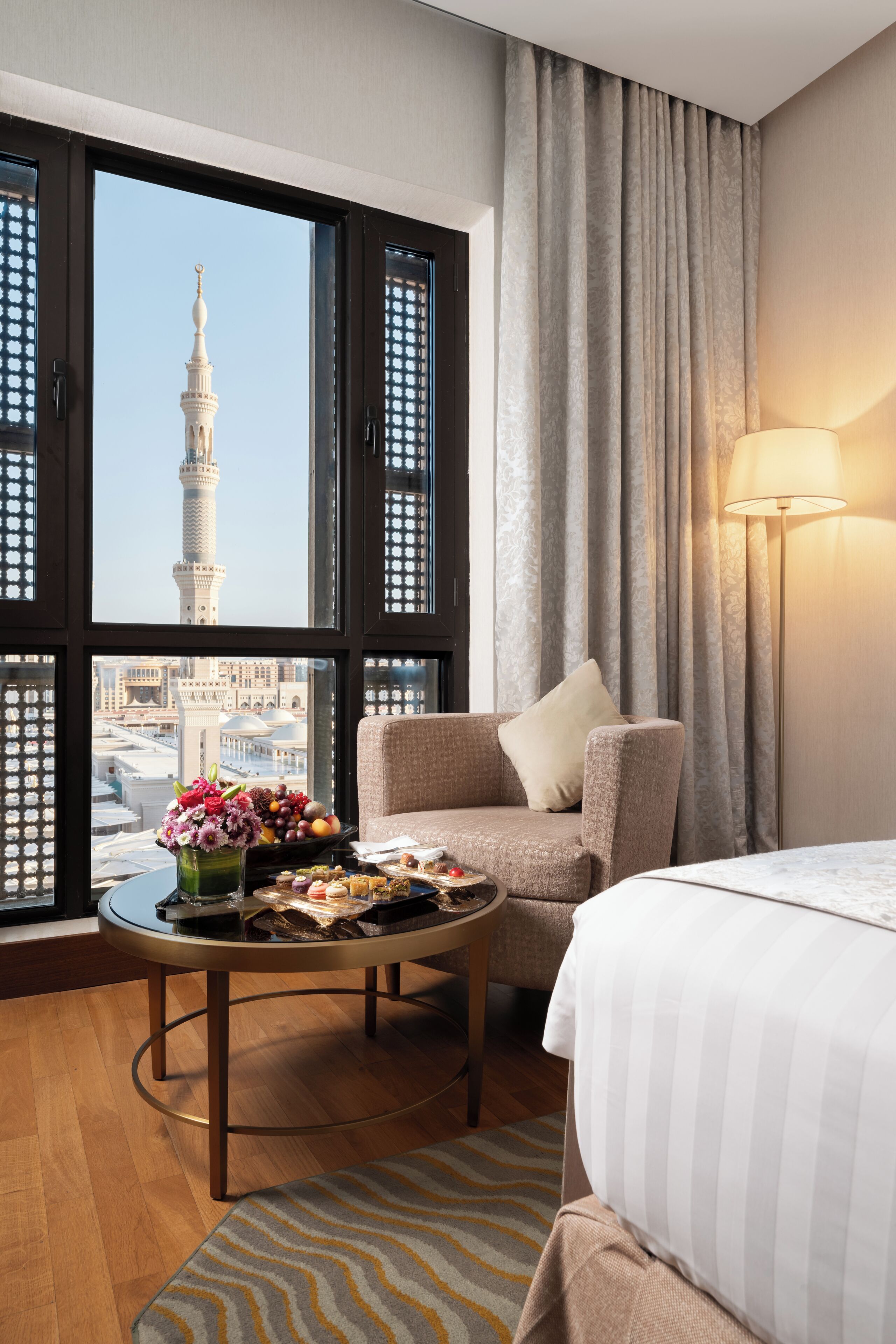 A hotel room with a view of a tower, a dining table with food, a sofa, a lamp, and a bed.