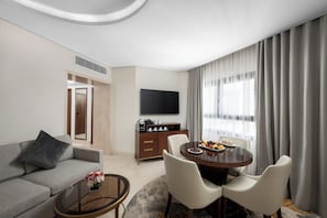 A modern hotel room with a sofa, a dining table with chairs, a coffee station, a TV, and a large window with curtains.