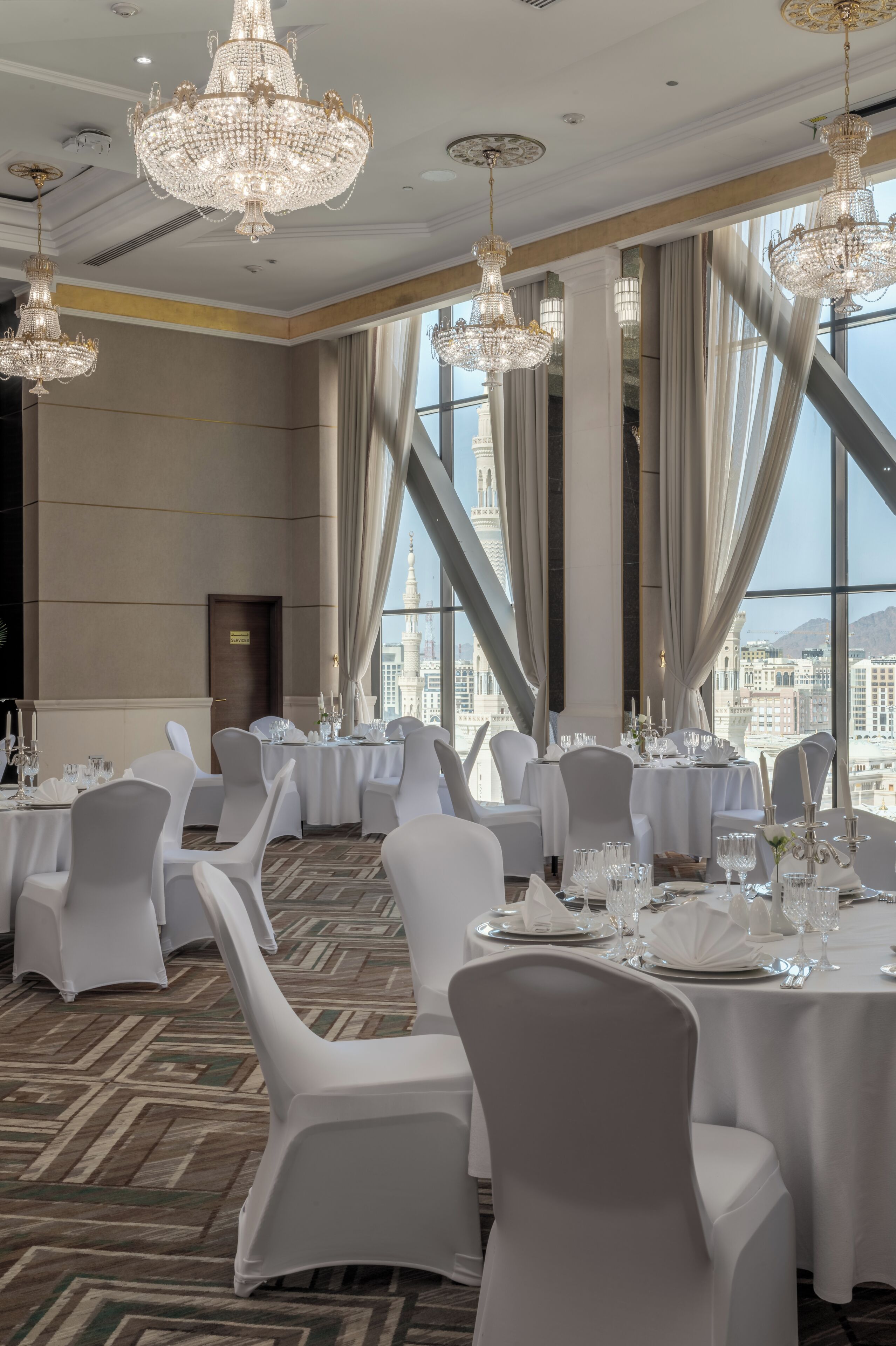 Un salón de banquetes con varias mesas dispuestas para comer, amplias ventanas que ofrecen vistas de la ciudad y lujosos candelabros.