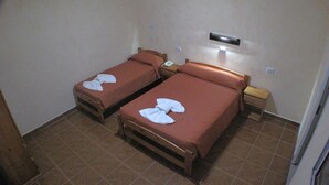 Una habitación con dos camas, cada una con una almohada y una toalla doblada. Entre las camas hay una mesita de noche con una lámpara.