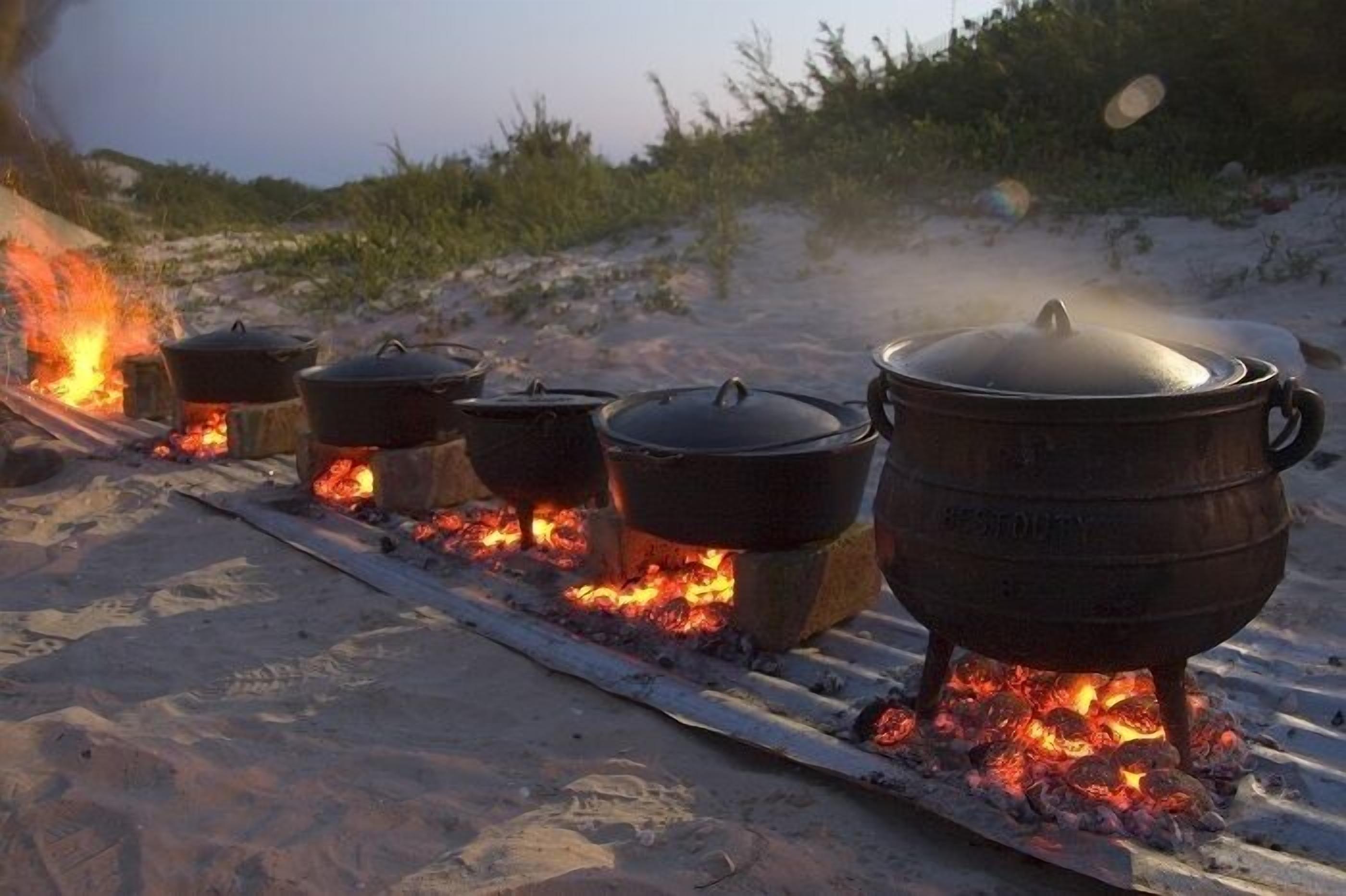 A row of pots over an open fire on a sandy surface.