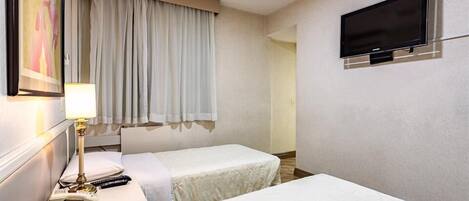 Una camera d'albergo con due letti, una televisione montata a parete, una finestra con tende e un comodino con lampada e telecomandi.