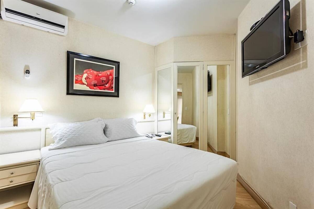 Un cuarto de hotel con una cama grande, un televisor de pantalla plana montado en la pared, una obra de arte enmarcada sobre la cama y un espejo en una esquina.