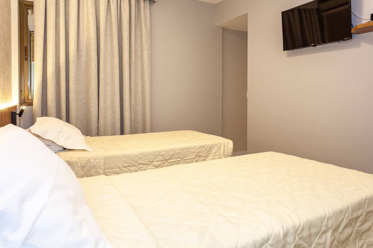 Habitación de hotel con dos camas, un televisor de pantalla plana montado en la pared y una ventana con cortinas.