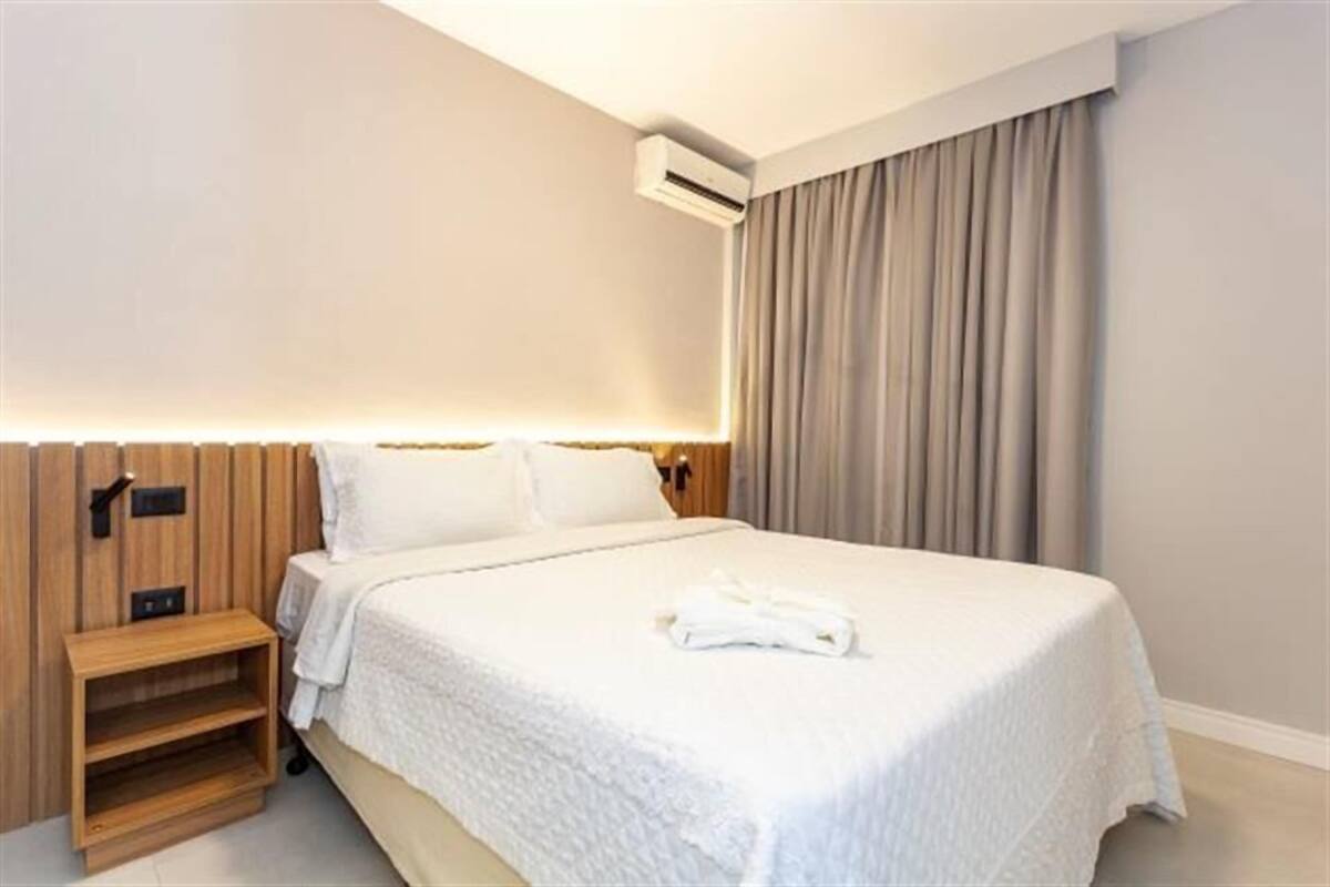 Una cama bien hecha con edredón blanco, una mesita de noche de madera y un equipo de aire acondicionado instalado en la pared.