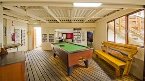 A pool table in a room with wooden flooring, a bench, and a staircase visible through a large window. - Island Vibe Knysna Hostel (Knysna)